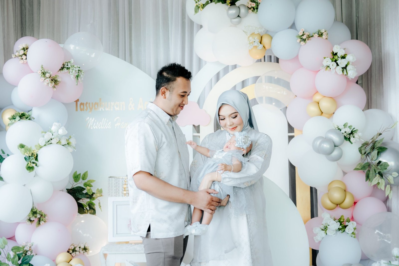 A family celebrating with pastel balloons, parents in elegant attire holding their baby.